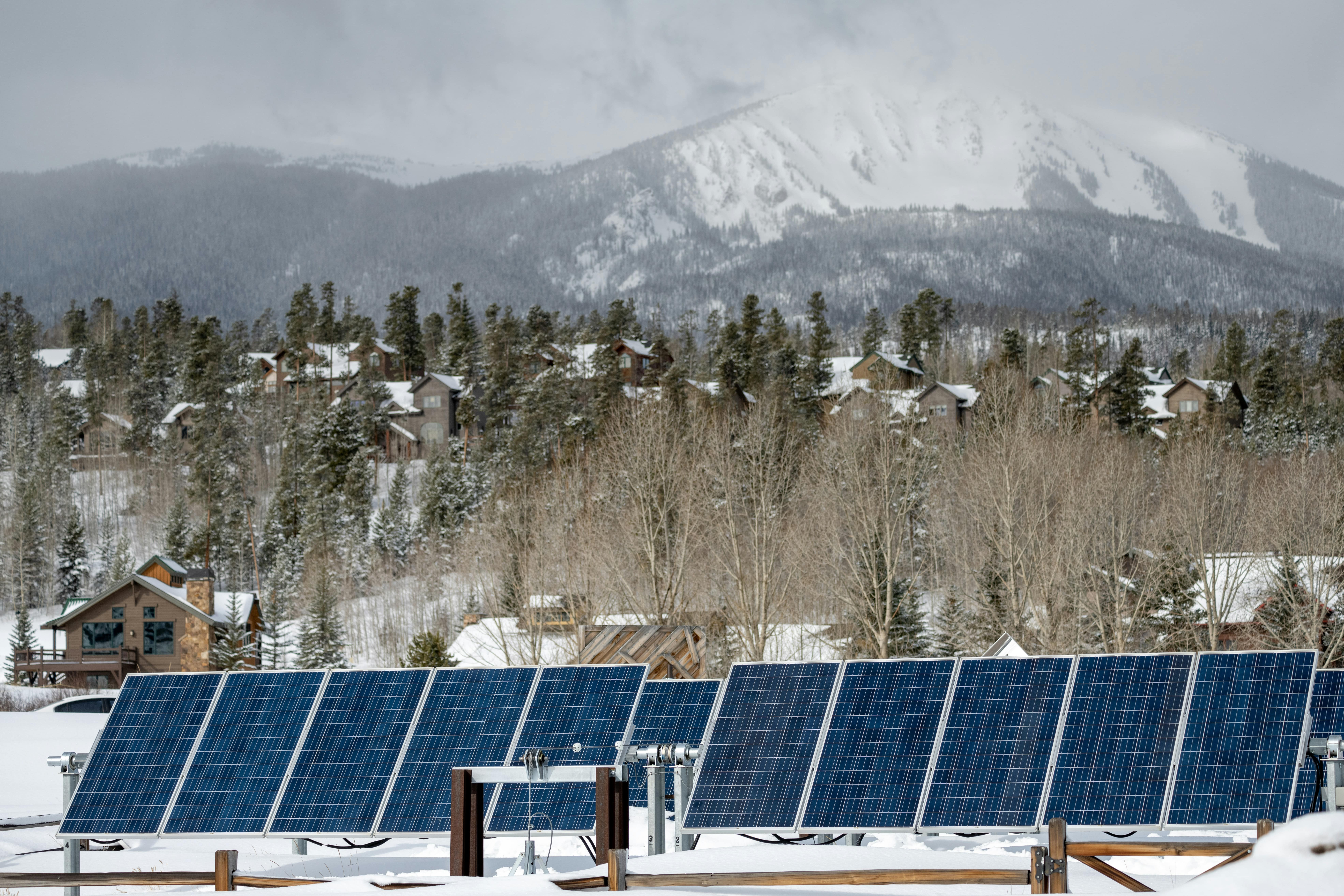 Solar panels covered in snow with a backdrop of a snowy forest and mountain, highlighting solar power in cold climates.